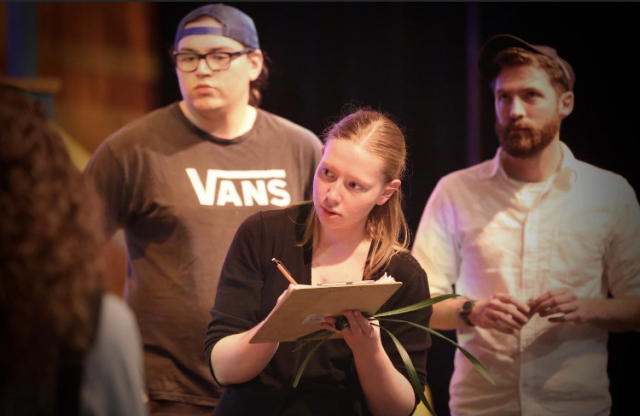 woman in the forefront of the stage writing on a clipboard with two men to the side.