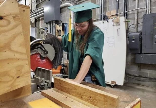 Person in a graduation outfit sawing a piece of wood.