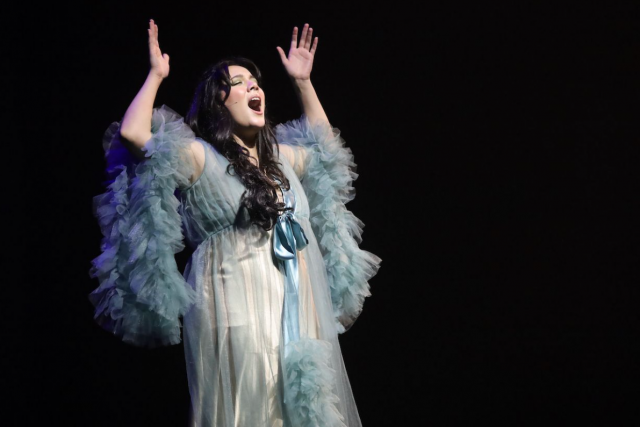 woman in light blue feathery and sheer dress singing infront of a spotlight